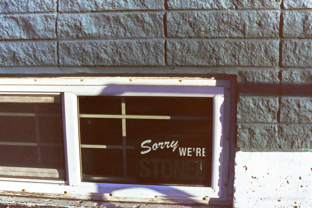 A window on a green brick building.  Barely readable in the window is a sign that reads 'sorry, we're stoned' in the style of a 'sorry, we're closed' sign.