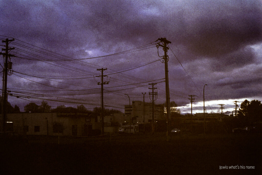 Photo of a cityscape beneath dramatic clouds.  Telegraph poles and power lines take up much of the photo.  The colours are washed out and faded, or else too dark, giving it the look of a photograph that has faded with time.